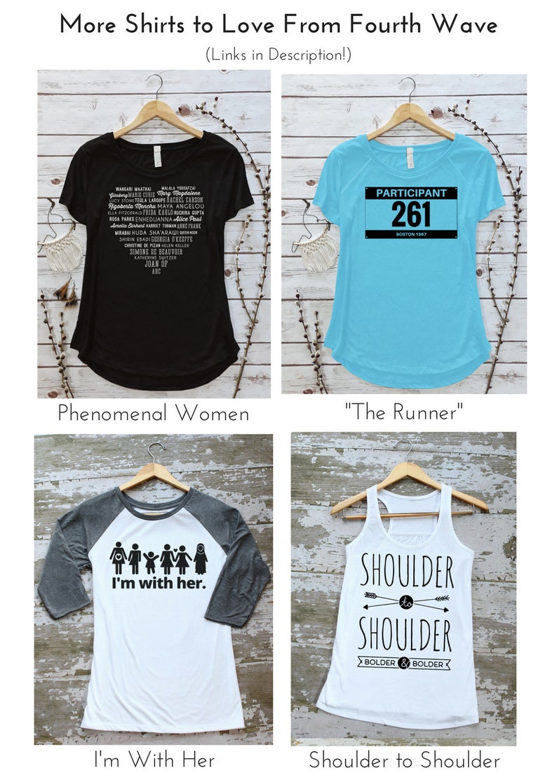 May include: Four different t-shirt designs on hangers. The first shirt is black with white text listing the names of many women. The second shirt is light blue with black text that says "Participant 261 Boston 1967". The third shirt is white with a gray sleeve and black text that says "I'm with her." and has a graphic of a family. The fourth shirt is white with black text that says "Shoulder to Shoulder Bolder & Bolder" and has a graphic of two arrows pointing towards each other.