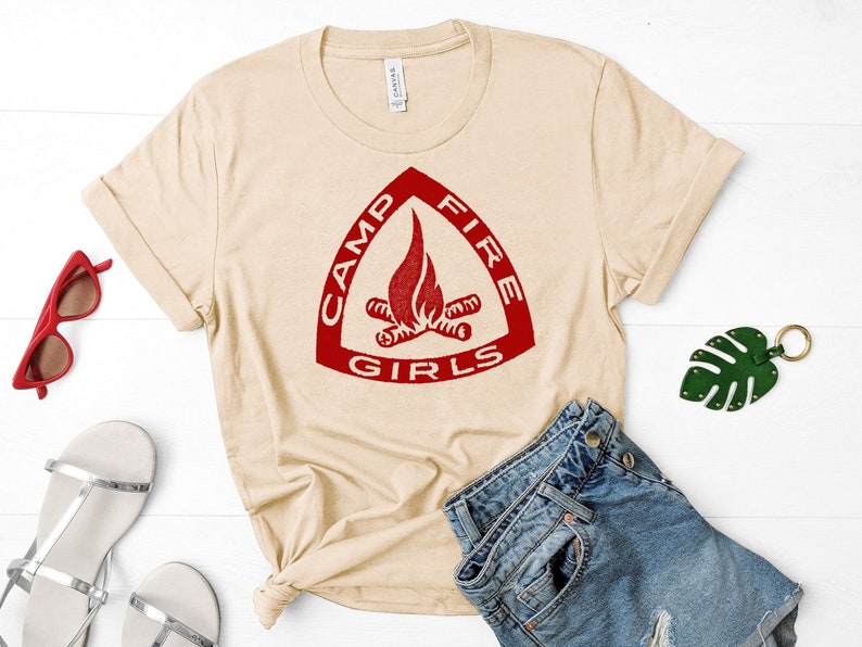 May include: A beige t-shirt with a red triangle logo that says "Camp Fire Girls" with a stylised image of a campfire inside the triangle.