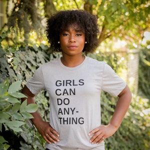 May include: A person wearing a grey t-shirt with the text "GIRLS CAN DO ANY-THING" printed on it. They are standing in front of a green leafy background.