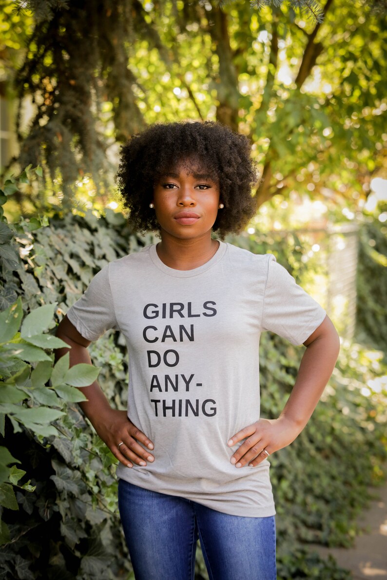 May include: A person wearing a grey t-shirt with the text "GIRLS CAN DO ANY-THING" printed on it. They are standing in front of a green leafy background.