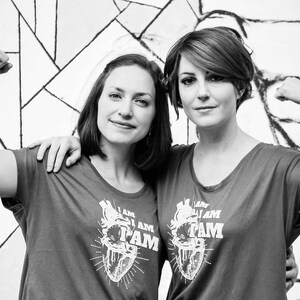 K&ouml;nnte beinhalten: Zwei Personen tragen graue T-Shirts mit einer stilisierten Herzgrafik und dem aufgedruckten Text "I am Fam". Beide beugen ihre Bizepse mit erhobenen Armen.