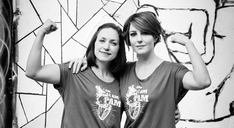 K&ouml;nnte beinhalten: Zwei Personen tragen graue T-Shirts mit einer stilisierten Herzgrafik und dem aufgedruckten Text "I am Fam". Beide beugen ihre Bizepse mit erhobenen Armen.