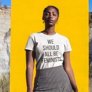 May include: A person wearing a white t-shirt with the text "WE SHOULD ALL BE FEMINISTS" printed on it. The person is standing in front of a yellow background.