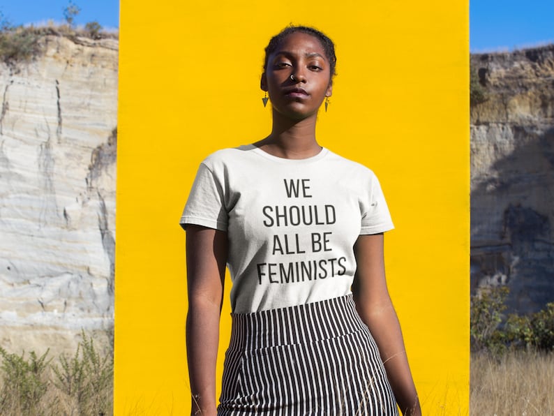 May include: A person wearing a white t-shirt with the text "WE SHOULD ALL BE FEMINISTS" printed on it. The person is standing in front of a yellow background.
