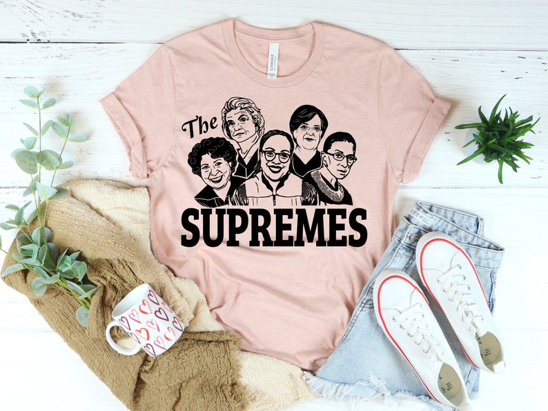 Pu&ograve; includere: Una maglietta rosa con una grafica nera di cinque volti di donne e il testo "The Supremes".