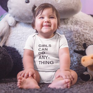 Pu&ograve; includere: Un bambino che indossa un body bianco con il testo "Girls can do anything" in nero. Il bambino &egrave; seduto su un tappeto soffice con una mucca di peluche grande e una mucca di peluche pi&ugrave; piccola accanto.