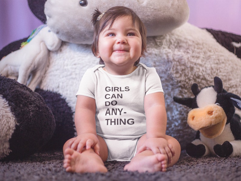 Pu&ograve; includere: Un bambino che indossa un body bianco con il testo "Girls can do anything" in nero. Il bambino &egrave; seduto su un tappeto soffice con una mucca di peluche grande e una mucca di peluche pi&ugrave; piccola accanto.