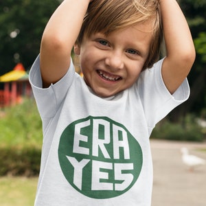 May include: A young child wearing a white t-shirt with a green circle and the text "ERA YES" in white letters.
