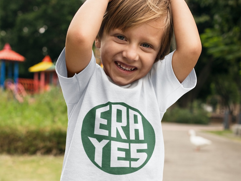 May include: A young child wearing a white t-shirt with a green circle and the text "ERA YES" in white letters.