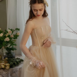 May include: A woman wearing a beige tulle wedding dress with a lace bodice and a floral crown. She is holding a bouquet of white flowers.