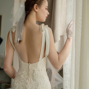 May include: A white lace wedding dress with a low back and a satin bow detail on the straps. The dress has a fitted silhouette and a long skirt. The model is wearing white gloves and a veil.