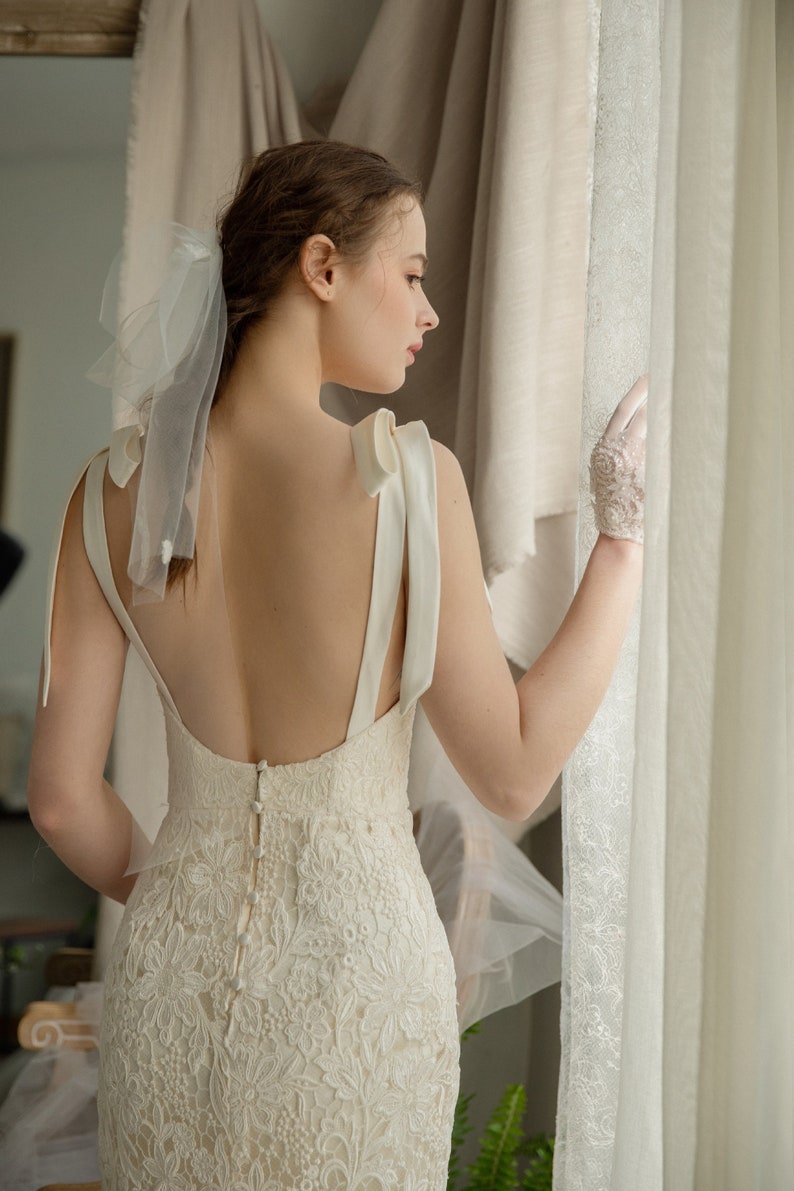 May include: A white lace wedding dress with a low back and a satin bow detail on the straps. The dress has a fitted silhouette and a long skirt. The model is wearing white gloves and a veil.