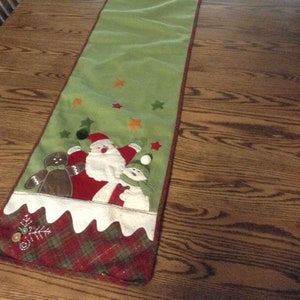 May include: Green Christmas table runner with Santa Claus, snowman, and gingerbread man appliqués. Features red, orange, and green star accents. The bottom edge has a red and green plaid pattern.