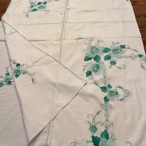 May include: A white rectangular tablecloth featuring embroidered floral patterns in shades of teal and green. The fabric displays decorative stitching along the seams and edges, with large flower motifs arranged in a corner-focused design.