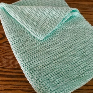 May include: A folded, light turquoise crochet blanket. The blanket is made with a textured stitch, and the edges are neatly finished. The soft, pastel color and handmade appearance suggest a cozy, comforting item, suitable for a baby or as a decorative throw.