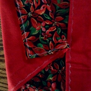 May include: A red fleece blanket with a poinsettia floral pattern in red, green, and white on a black background. The edges are finished with white stitching.