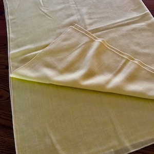 May include: A folded, pale yellow linen tablecloth. The fabric has a subtle texture and is partially folded over, revealing the underside. The tablecloth is on a dark wooden surface.