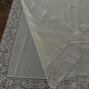 May include: A white rectangular tablecloth featuring a smooth central fabric panel bordered by a wide, intricate floral lace trim. The corner of the cloth is folded back to reveal the underside, showcasing the detailed lace pattern and fabric texture.