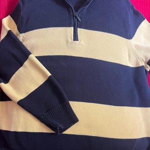 May include: A long-sleeve quarter-zip sweater featuring wide horizontal stripes in navy blue and beige. The garment has a ribbed collar, cuffs, and hem, with a Villager brand label visible at the inner neckline. The zipper pull is navy blue.