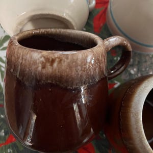 May include: A collection of ceramic mugs and a small bowl. The primary mug is brown with a lighter brown rim. Other mugs are cream-colored with pink and blue stripes. The items are on a patterned surface.