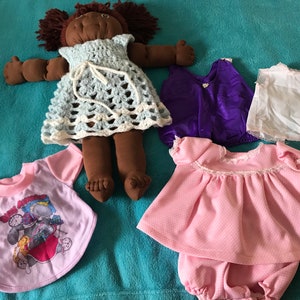 May include: A brown rag doll with black hair wearing a crocheted blue and white dress. The doll is surrounded by four different outfits for the doll, including a pink dress, a purple sleeveless top, a white sleeveless top, and a purple and pink t-shirt.