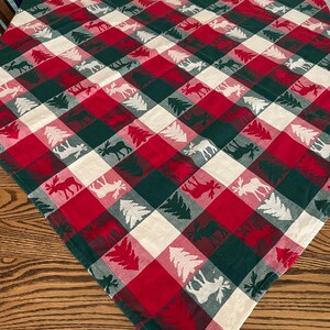 May include: A red, green, and white plaid tablecloth with a festive design. The pattern features alternating squares of red, green, and white, with silhouettes of moose and pine trees. The tablecloth is made of a soft fabric and is suitable for a holiday gathering.