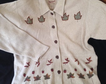 May include: A beige cardigan with a button-up front and long sleeves. The cardigan has a brown and green leaf design on the front and bottom. The cardigan has the text "Woolrich" on the collar.