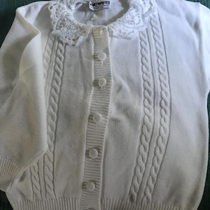 May include: White knitted cardigan with a white crocheted collar and button closure. The cardigan has a cable knit pattern and the label reads "Knitwaves".