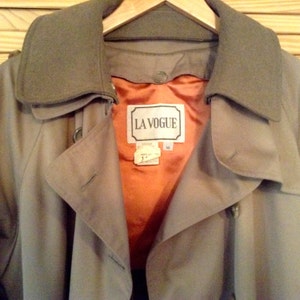 May include: A beige trench coat with a brown collar and a label that reads "LA VOGUE". The coat has a double-breasted closure and a belt.