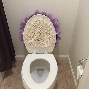 May include: A white toilet with a purple crocheted toilet seat cover. The cover has a white crocheted doily in the centre.