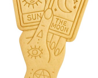 May include: A light brown cookie in the shape of a hand holding two tarot cards. The cards read "SUN" and "THE MOON" with sun and moon illustrations. The hand has an eye, triangle, and pentagram design.