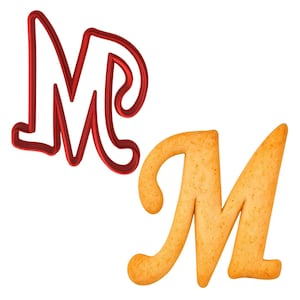 May include: A red metal letter "M" cookie cutter next to a baked letter "M" cookie. The cookie cutter is a shiny, solid red. The baked cookie is golden brown with a slightly textured surface, and is in the shape of the letter "M".