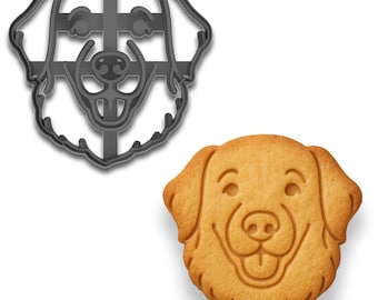 May include: A gray metal dog-shaped cookie cutter next to a baked golden-brown cookie in the shape of a dog's face. The cookie has etched details for the eyes, nose, and mouth, creating a smiling expression. The cutter is a silhouette of a dog's head.