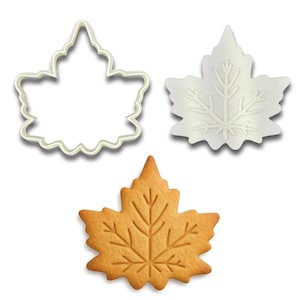 May include: Three maple leaf-shaped items: a cookie cutter, a white leaf-shaped item with etched details, and a baked cookie. The cookie cutter is white with a scalloped edge. The cookie is golden brown with leaf vein details.