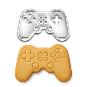 May include: A silver metal cookie cutter in the shape of a video game controller is positioned above a baked cookie in the same shape. The cookie is golden brown with detailed button impressions.