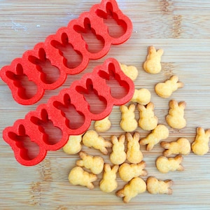 May include: Red plastic bunny-shaped cookie cutters next to several bunny-shaped crackers. The cutters are arranged in rows of five bunny shapes. The crackers are golden yellow and scattered around the cutters on a wooden surface.