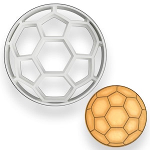 May include: A white plastic soccer ball-shaped cookie cutter next to a golden-brown soccer ball. The cookie cutter has a geometric design with pentagons and hexagons. The golden-brown soccer ball has a similar geometric pattern.