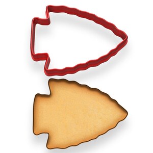 May include: A red plastic arrowhead-shaped cookie cutter sits above a baked cookie in the same shape. The cookie has a light brown color and a slightly textured surface. The cutter is a solid, vibrant red.