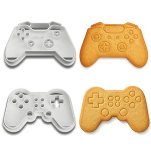 May include: Four items: two white plastic cookie cutters and two baked cookies, all in the shape of video game controllers. The cookies are golden brown with detailed button impressions. The cutters are white.
