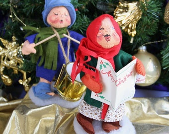 Könnte beinhalten: Zwei handgefertigte Weihnachtsfiguren. Eine Figur spielt eine Trommel, trägt ein blaues Outfit und einen Hut mit einem grünen Schal. Die andere Figur trägt einen roten Schal und einen grünen Mantel und hält eine Karte mit "Merry Christmas".