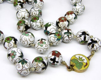 May include: A close-up of a necklace with white, round beads. Each bead is decorated with a floral design in red, green, and black, with gold accents. The necklace is strung on a beige cord and has a yellow clasp.