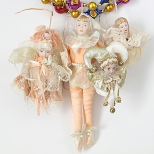 May include: A collection of vintage Christmas ornaments featuring porcelain doll faces and delicate fabric costumes. The ornaments include an angel, a jester, and a lady, all in soft pastel colors with lace and pearl accents. They hang from a colorful beaded garland.