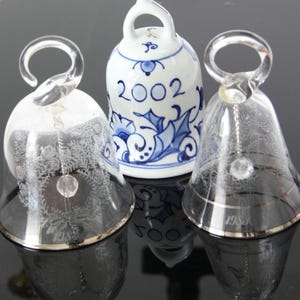 May include: Three decorative bells. Two are clear glass with silver rims and etched designs, one with the year 1988. The third is white ceramic with blue floral patterns and the year 2002. All have clear glass handles.