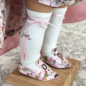 May include: A pair of white knee-high socks with pink ribbon ties and embroidered details. The socks are worn with pink floral print shoes with a buckle closure.