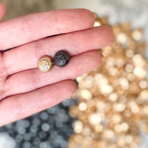May include: Close-up of two decorative buttons. One button is gold-colored, the other is dark gray. Both buttons have a detailed embossed design. The background is blurred with more buttons in gold and dark gray.