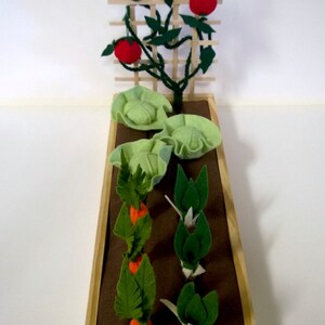 May include: A wooden planter box with brown felt lining. The planter contains felt representations of a tomato plant with red tomatoes, two heads of cabbage, and three rows of carrots.