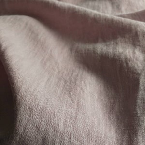 Pode incluir: Um close-up de um tecido de linho macio, rosa claro, com uma textura sutil. O tecido est&aacute; dobrado e drapeado, mostrando seu caimento e textura.