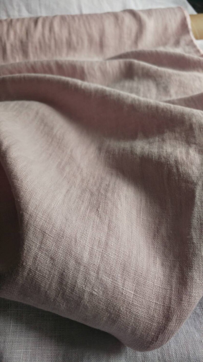 Pode incluir: Um close-up de um tecido de linho macio, rosa claro, com uma textura sutil. O tecido est&aacute; dobrado e drapeado, mostrando seu caimento e textura.