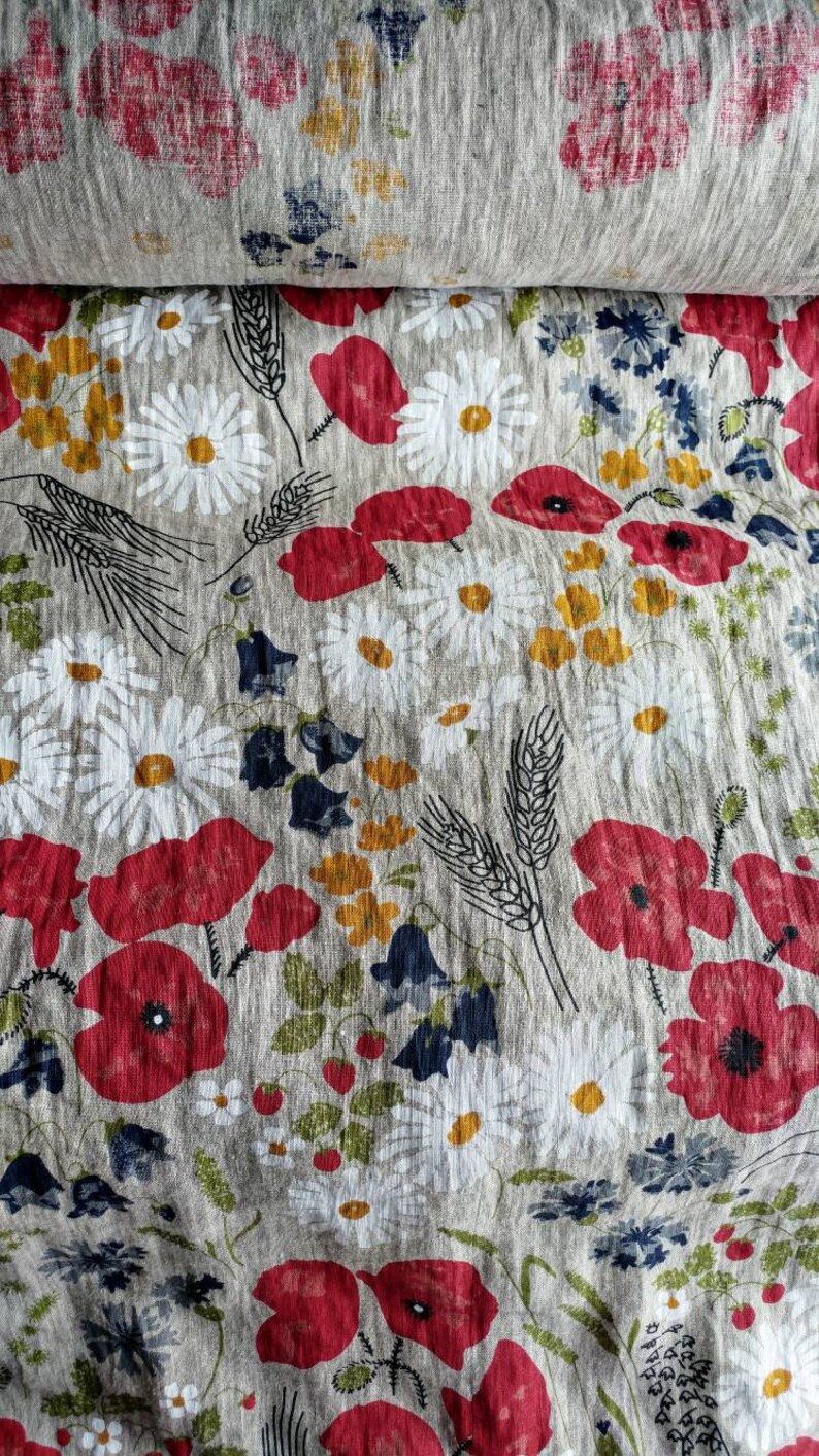 May include: A close-up of a fabric with a floral pattern. The fabric is a light beige color with red poppies, white daisies, and green leaves. The fabric has a textured, linen-like appearance.