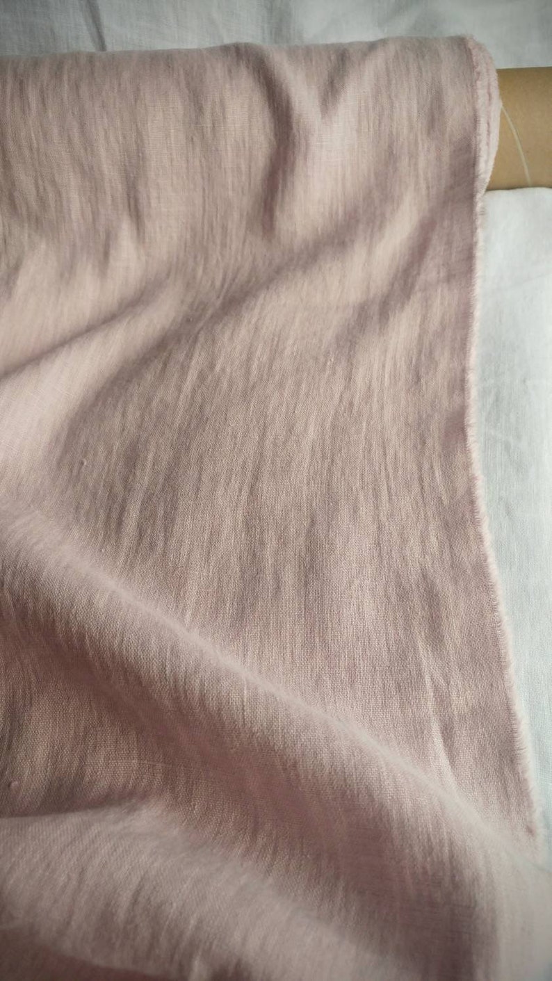 Pode incluir: Um close-up de um tecido de linho macio de cor rosa claro. O tecido est&aacute; dobrado e drapeado, mostrando sua textura e caimento.